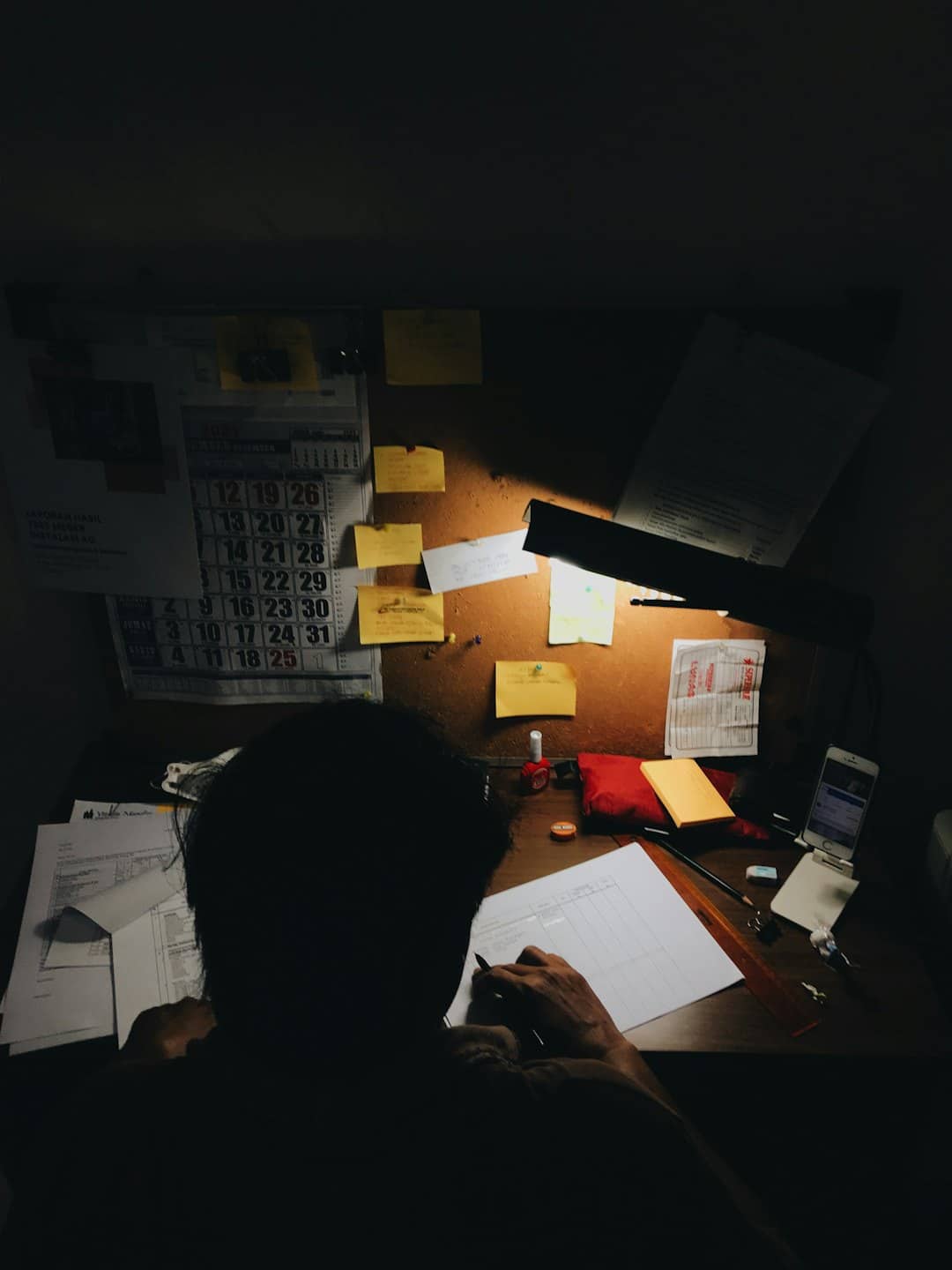 Silhouettenhafte Person schreibt an einem unordentlichen Schreibtisch unter einer Lampe.
